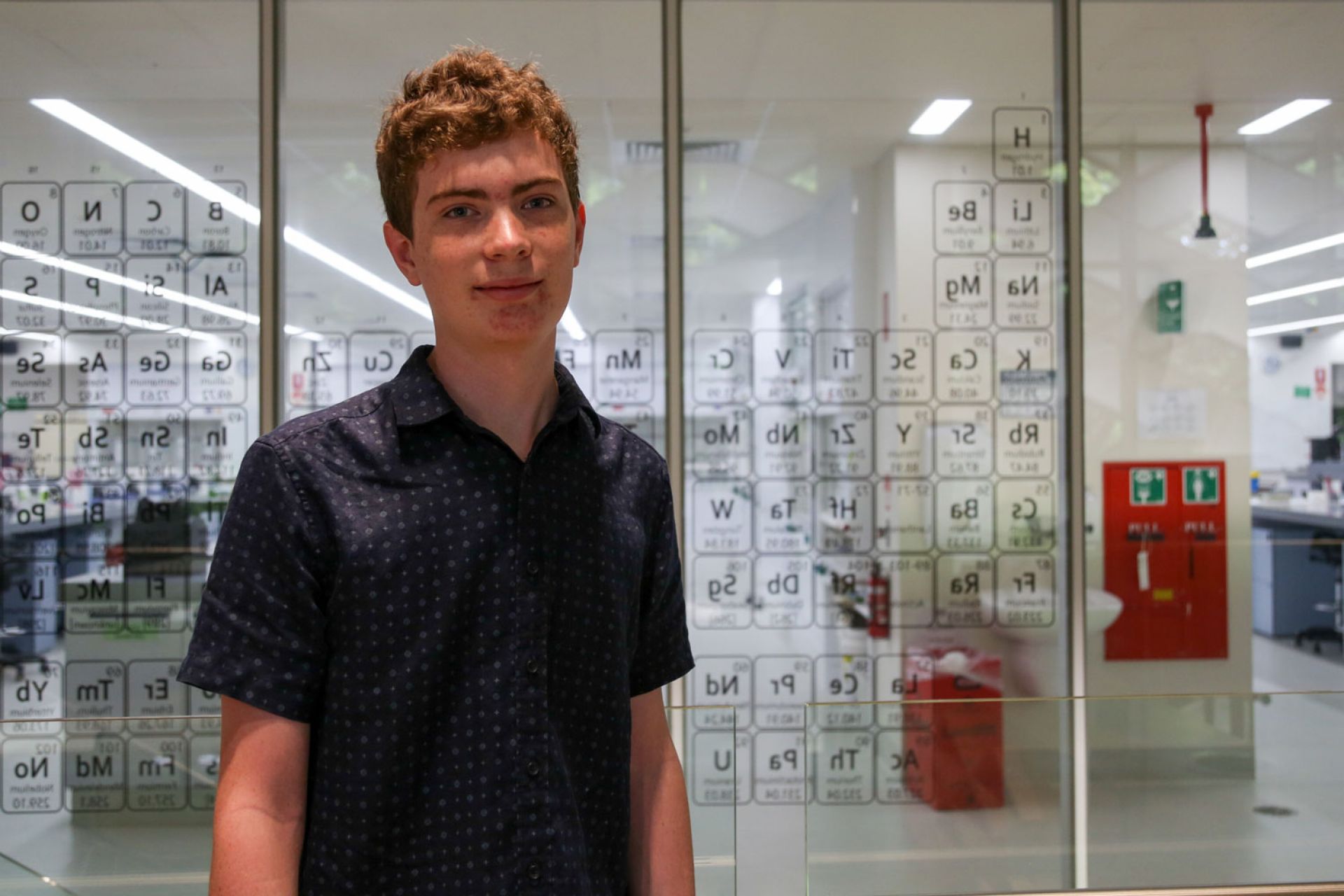 Portrait of Patrick, periodic table design on a window is behind him, the window leads to a lab