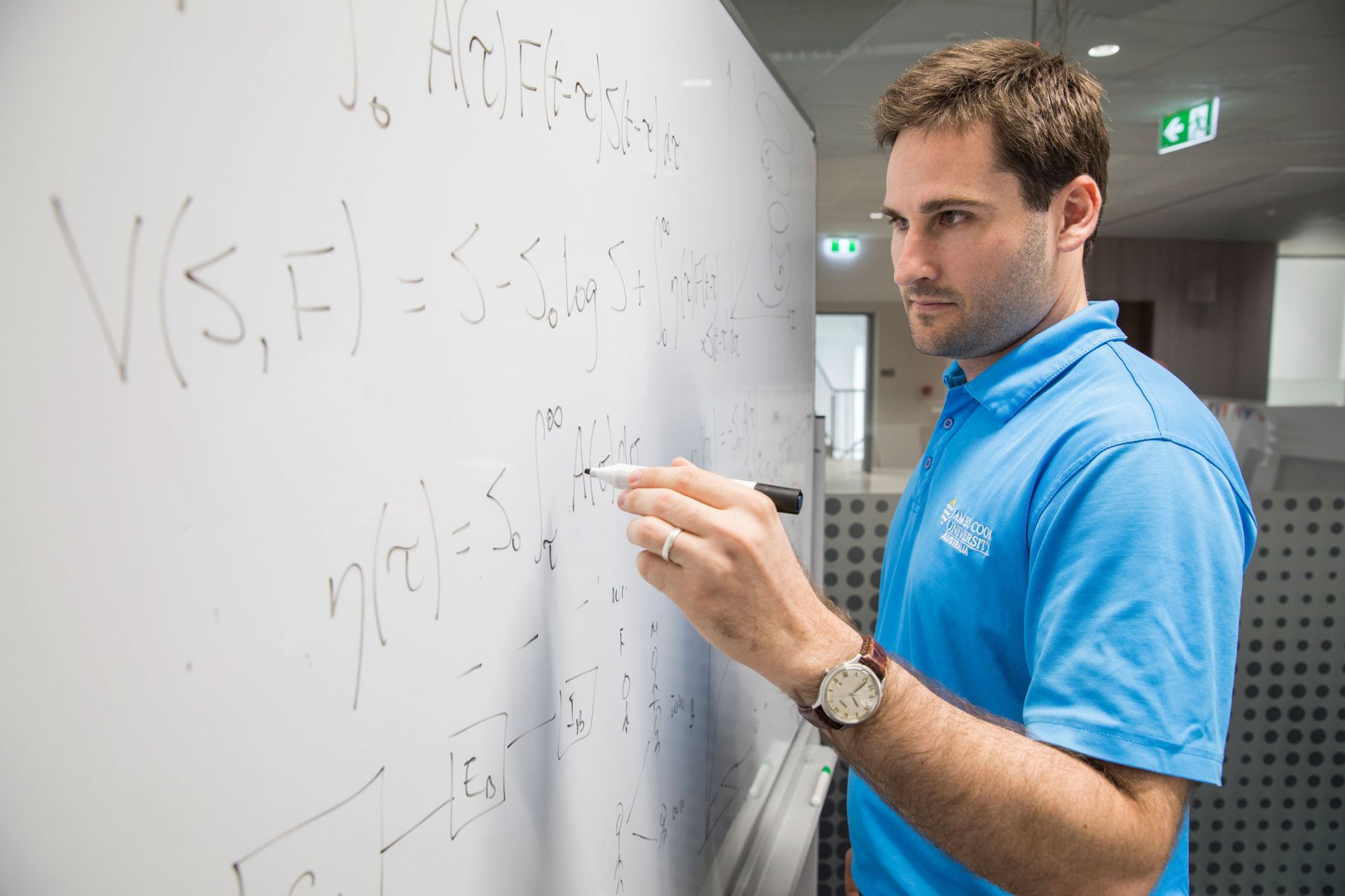 A man in a blue shirt is side-on to a white board where he's writing equations