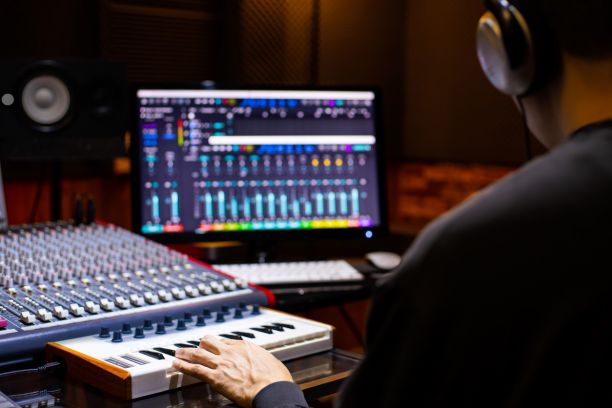 The back of a person wearing headphones and sitting in a sound recording studio with a keyboard and sound board. 