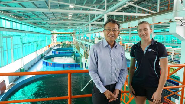 JCU PhD Candidate Phoebe Arbon smiling and standing next to an aquaculture researcher in Singapore. In the background are many prawn aquaculture tanks. 