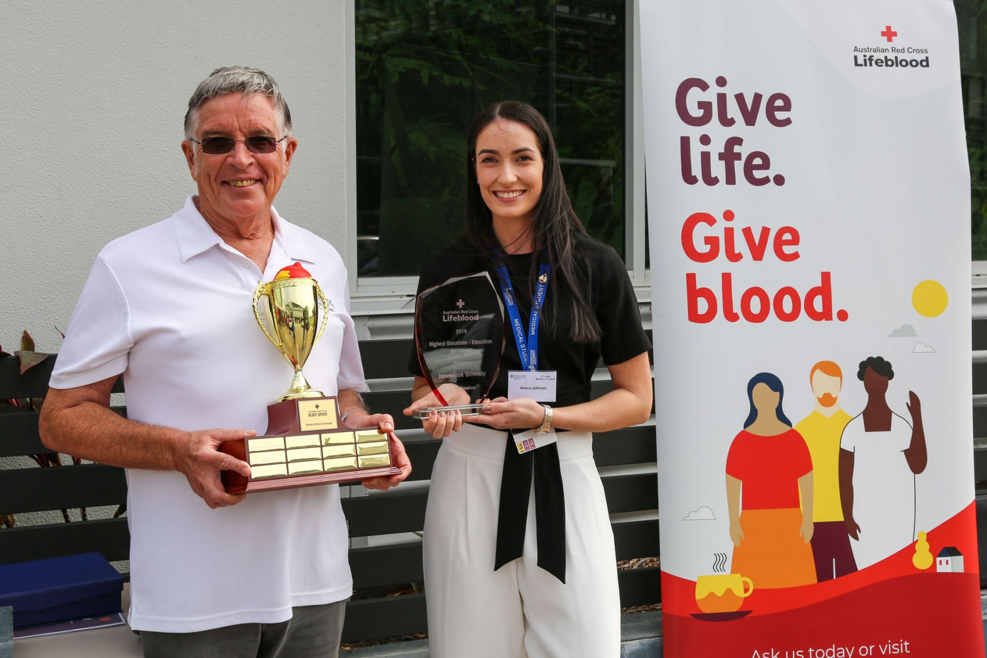 Bill Tweddell and Bianca Johnson hold trophies and stand next to a blood donation promotional banner