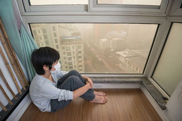 Child inside a highrise apartment wearing a mask and looking at a smog filled city