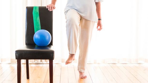 The legs of a lady wearing cream pants and balancing on one leg while holding onto a black chair with a small exercise ball and resistance band on it. 