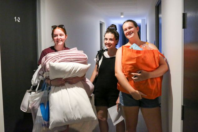 Three female students, two holding bags and linen, smile for the camera in a corridor. 