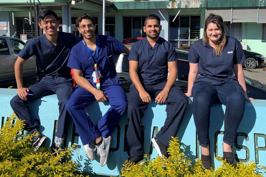 Four students wearing dark blue scrubs sit on a bright blue wall in front of a building