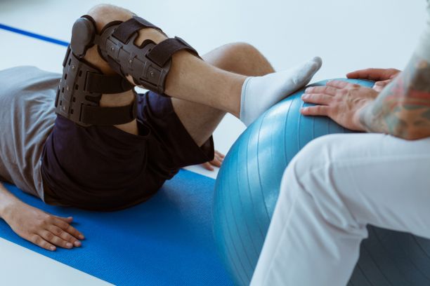 A person with a leg brace on is laying on their back on a yoga mat with their right foot on a yoga ball that someone else is holding steady. 