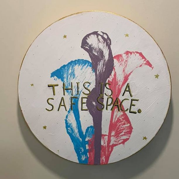 A circular painting with a white background and three flowers each in blue purple and red and has the words This is a safe space. 