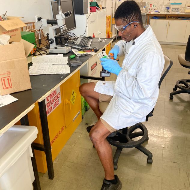 JCU PhD Candidate Emenike Okonkwo sitting on a chair working with a chemical solution in a beaker in a science laboratory. 
