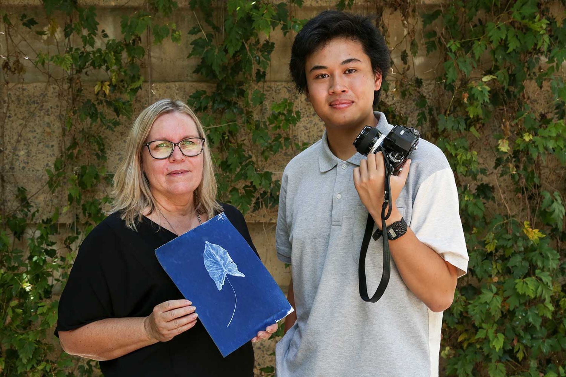 Ann holds a vibrant blue cyanotype image of a leaf, next to her is a young male holding a film camera