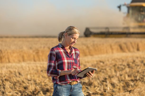 A women wearing a red check shirt and jeans looks at a digital tablet while grains are harvested on a farm behind her. 