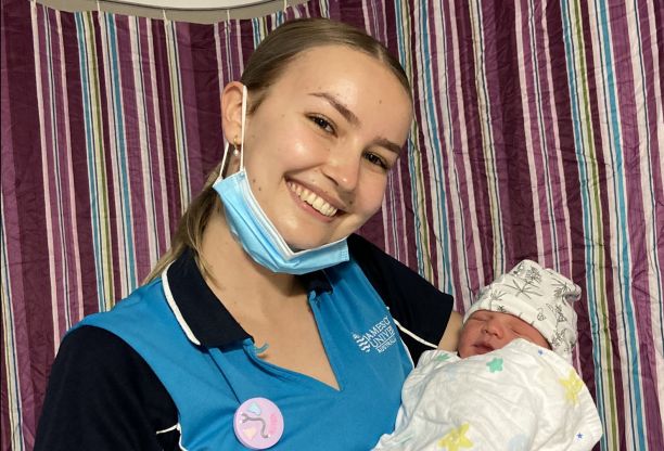 Kira holding a newborn while wearing midwifery student uniform. 