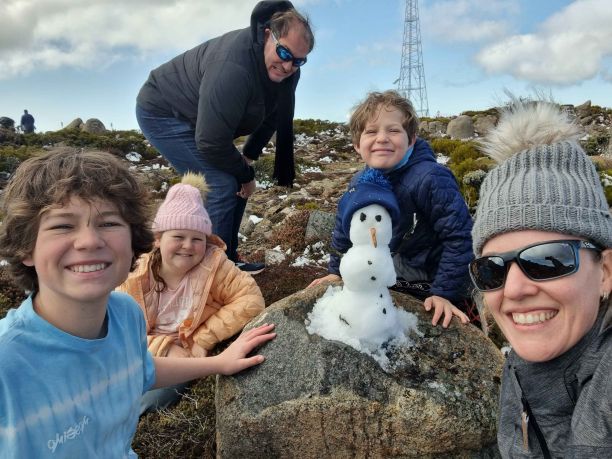Renee wearing a beanie and posing with her three children and husband in the snow. There is a snow man on a rock in the centre, with Renee and her family posing around it. 