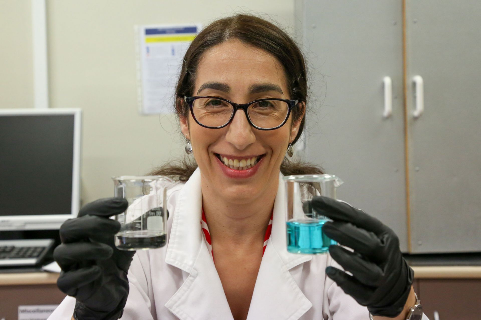 Close up photo of a female scientist in a lab coat holding two beakers, one with blue liquid and one with clear liquid