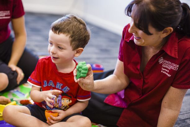 Female JCU speech pathologist wearing a red shirt holds up a plastic broccoli to a young boy sitting on the ground.