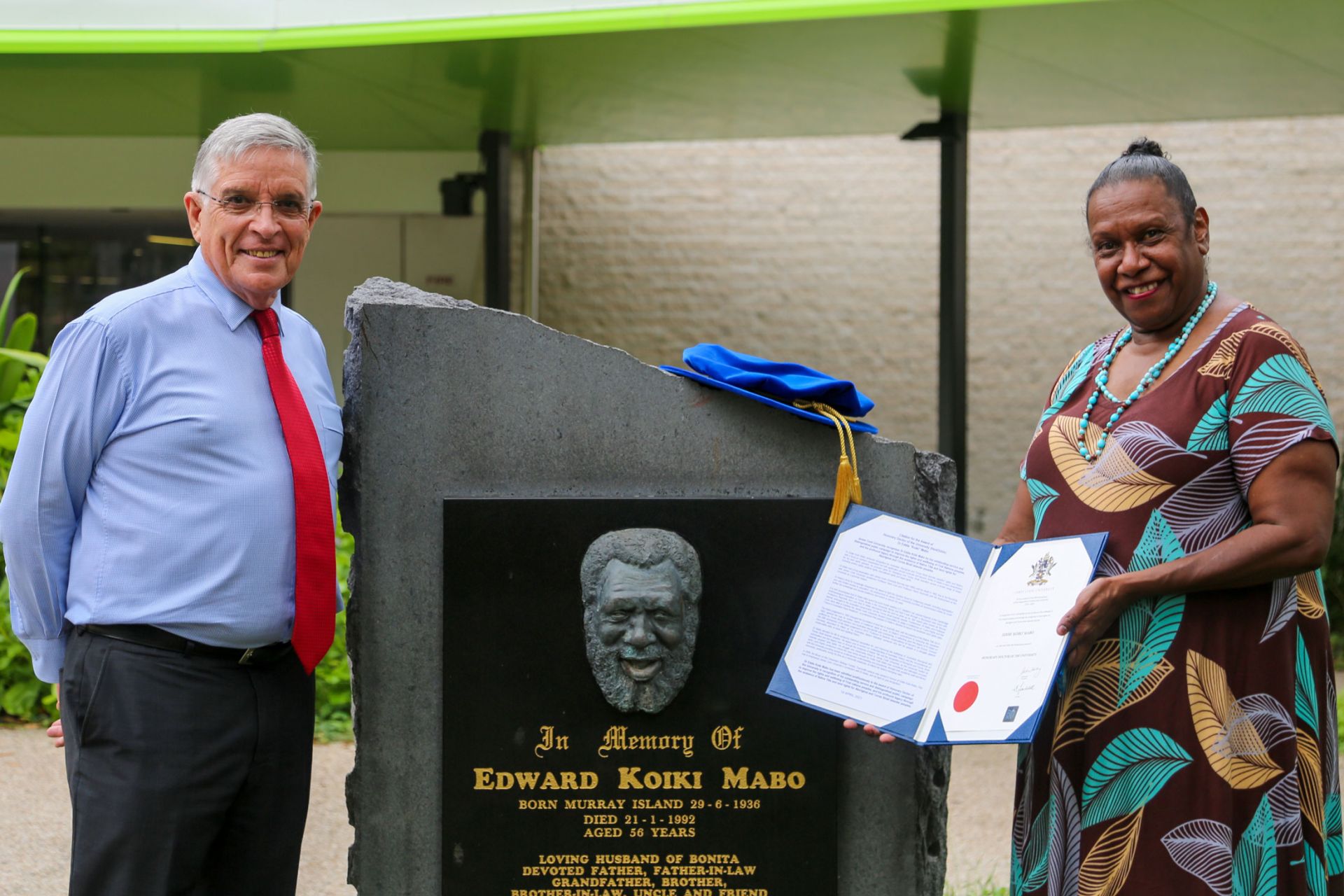 Chancellor Bill Tweddell and Gail Mabo stand on either side of a plaque commemorating Eddie. A PhD bonnet is on top of the plaque and Gail holds open a blue folder showing the testator