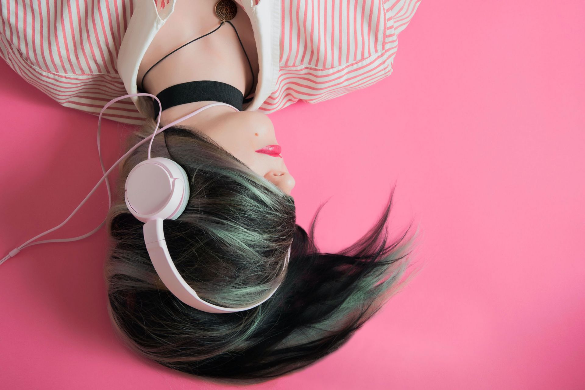 Woman lying down, pink background, hair covering her face and a pair of white headphones on