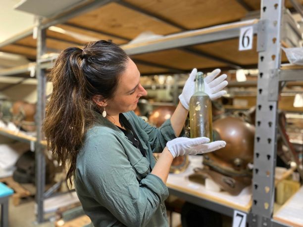 Maritime archaeologist Dr Maddy McAllister holds up an old bottle architect. In the background are shelves with other various maritime artifacts. 