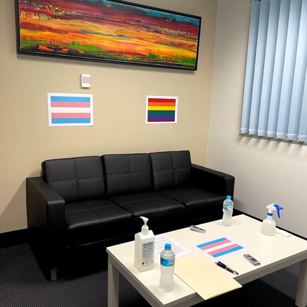 A room with a black lounge chair and a transgender and LGBTQI flag behind it and on the white coffee table in front of the lounge. 