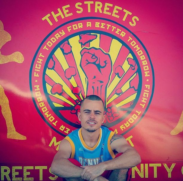 Jesse sitting on the ground in front of a sign for his first organisation, The Streets Movement, with his knees tucked to his chest.