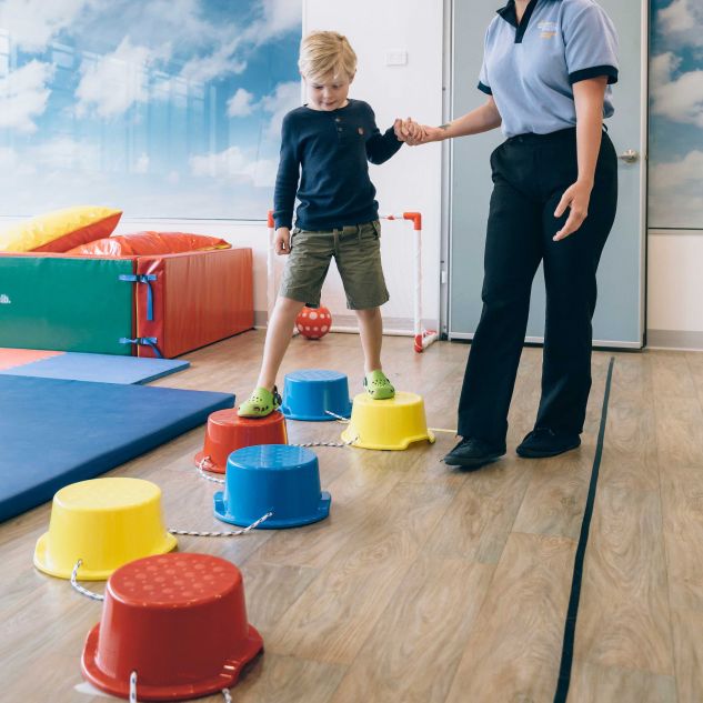 A young child balances on coloured buckets while holding the hand of a JCU occupational therapist wearing a light blue shirt and black pants. 