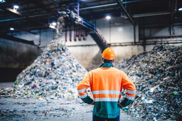 A person wearing fluro work clothes standing facing a large pile of waste at a recycling plant. 