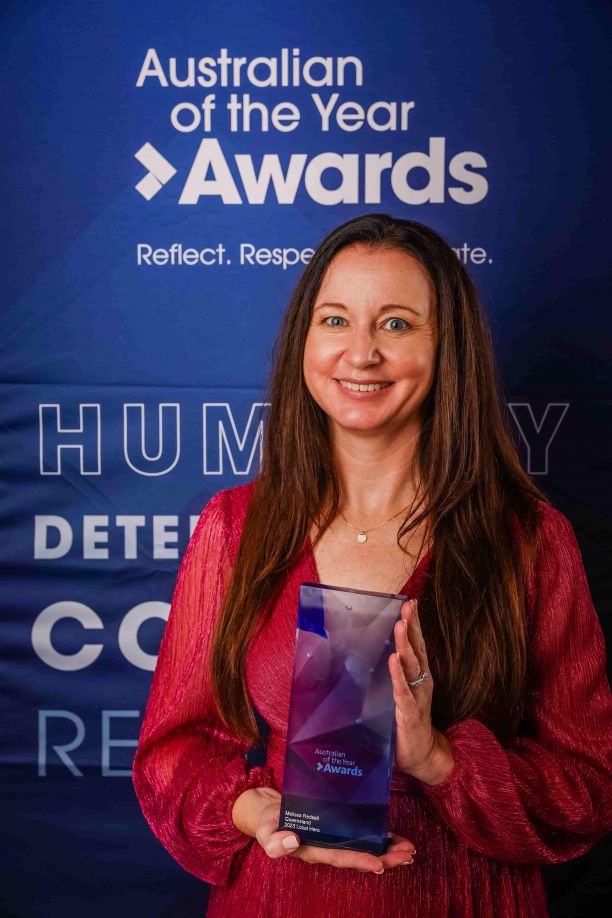 JCU Alumni Melissa Redsell smiling wearing a read dress and holding a blue glass trophy in front of a sign reading Australian of the year awards. 