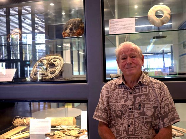 Laurie Bragge stands in front of a glass exhibition case holding pieces of his collection. 