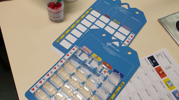 A weekly pharmacy blister pack lies on a table, with a list of instructions on how to fill it. There is a container of M&Ms in the top left corner. 