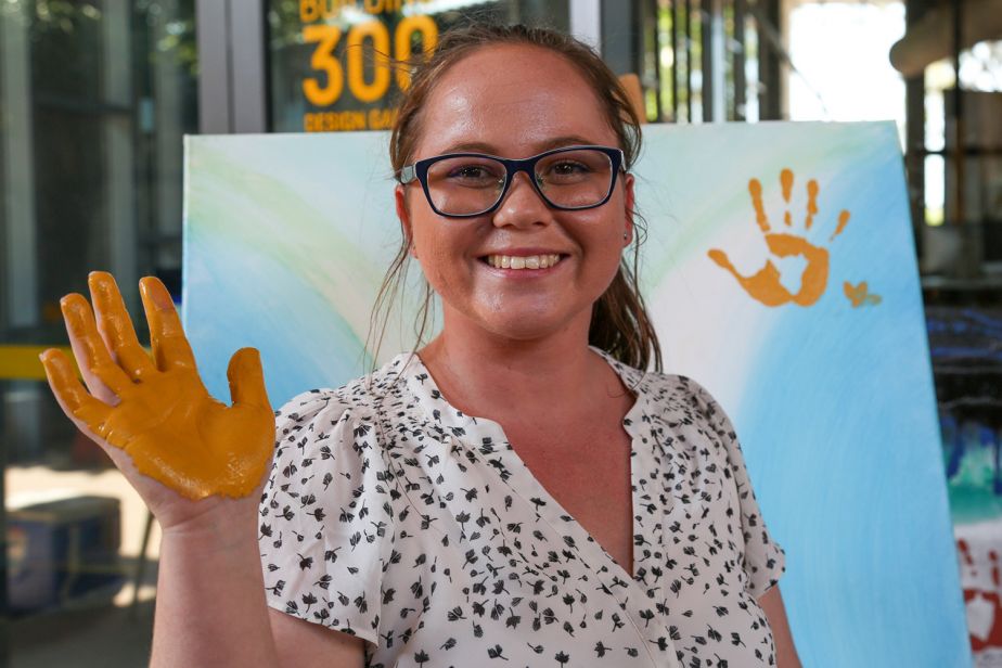 Nikitta smiles at the camera, holding up her right hand with the palm covered in yellow paint. behind her is a canvas with her hand print on it
