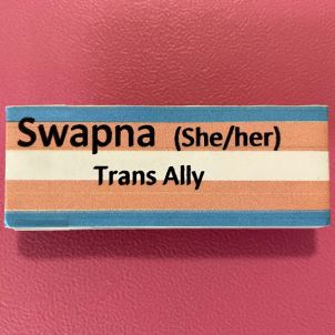 A name badge on a pink background that reads Swapna she / her and trans ally with the badge having blue pink and white stripes. 