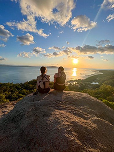 Two female students sitting on top of a hill overlooking the ocean at sunset