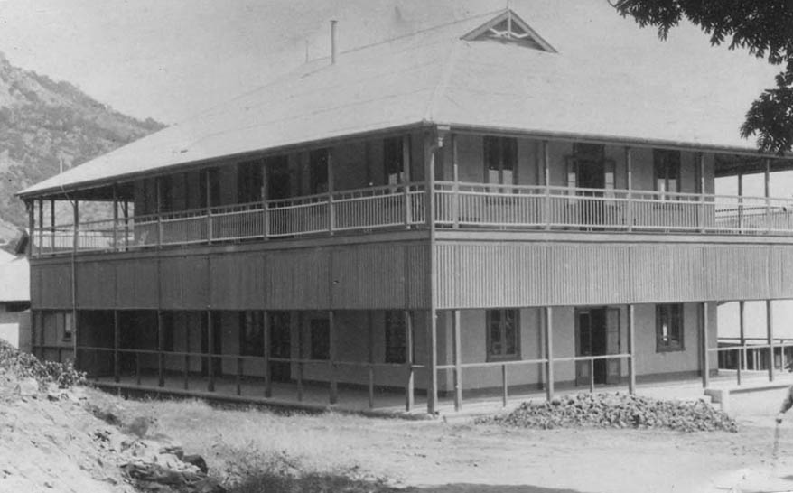 A black and white image of a large historical building, with two stories and a long, winding veranda. 