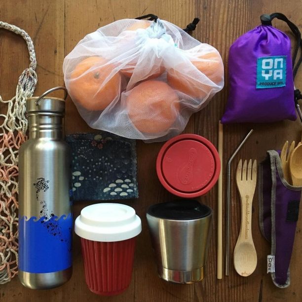 A reusable shopping kit laid out on a wooden table, with a woven shopping bag, metal water bottle, reusable coffee cup and cutler, and a bag of mandarines in a reusable produce bag. 