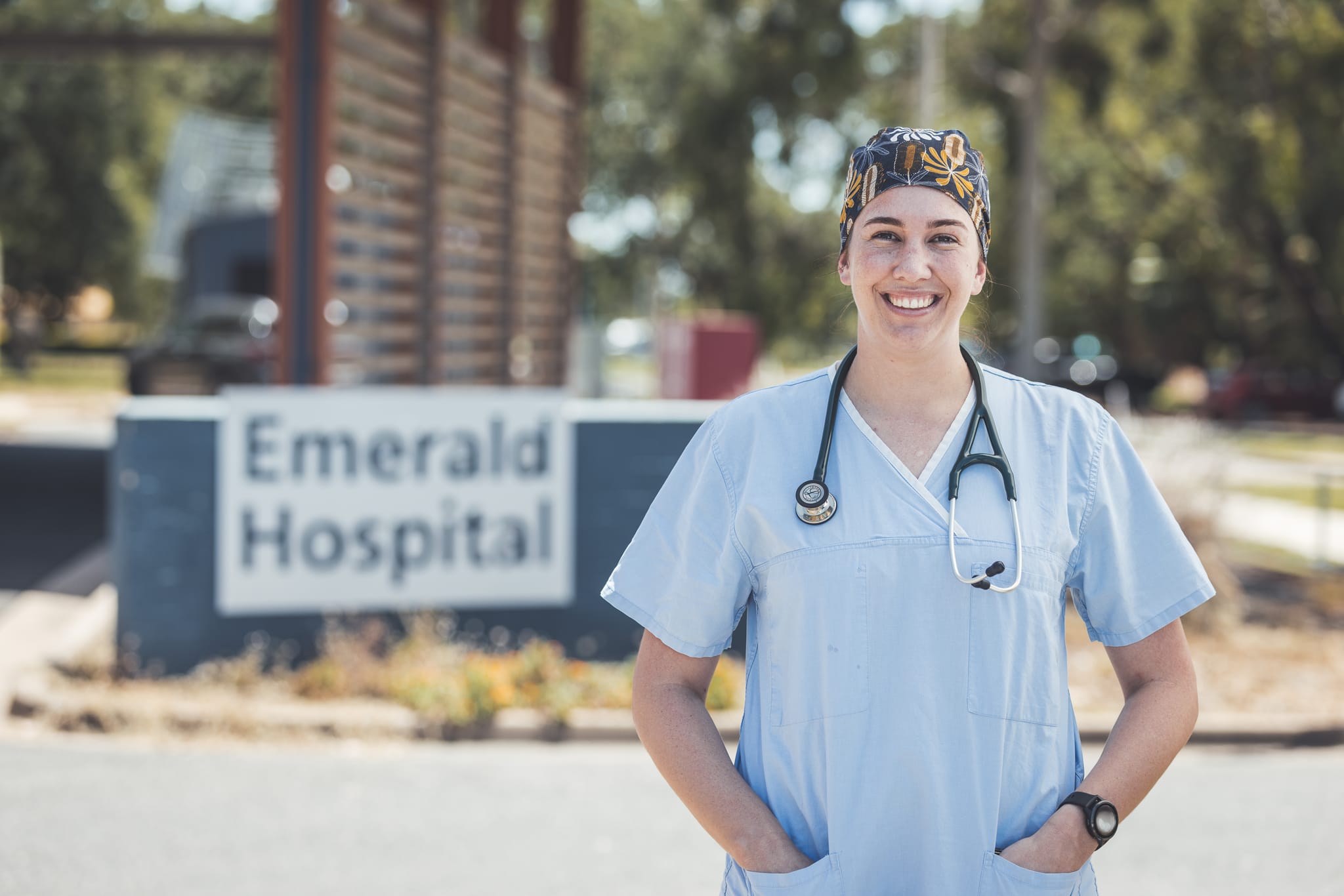 A female medical worker dressed in scrubs and wearing a stethoscope standing outside a sign for Emerald Hospital. 