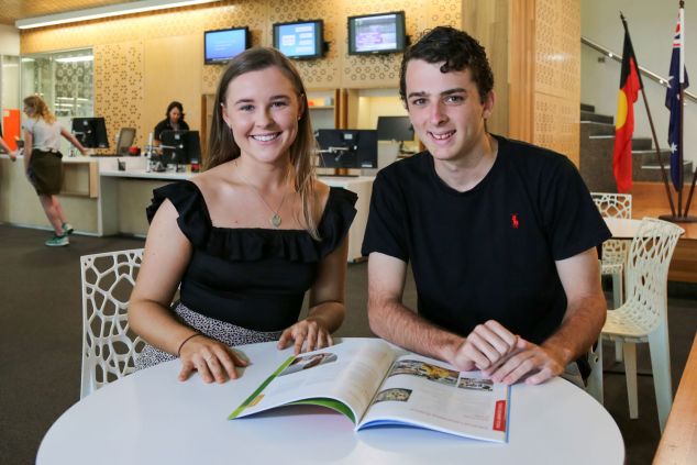 Sophie Horlock and William Thomas sit at a table in front of the student services desk.