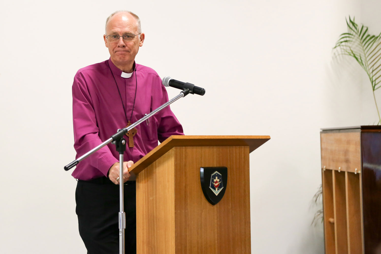 A man in a purple shirt and religious collar stands at a lectern
