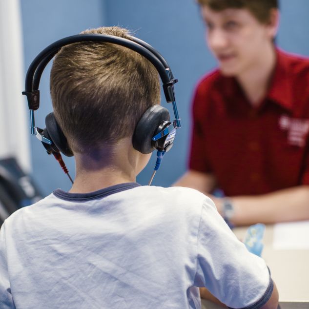 The back of a young boy sitting at a desk wearing a white shirt and over the ear headphones with a JCU speech pathologist wearing a red shirt in the background.