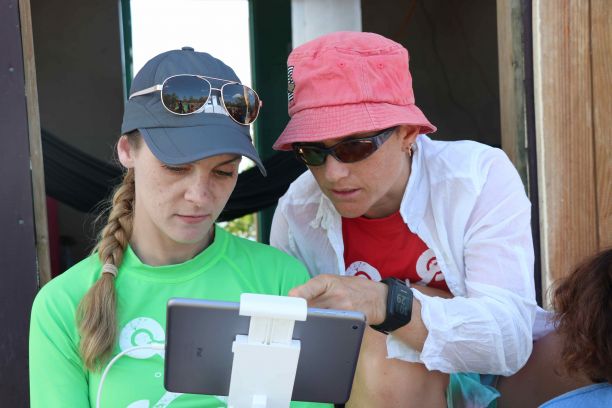 A woman wearing a blue cap and green rash shirt looks at an ipad with drone controls while JCU researcher Karen Joyce is on the right pointing to the screen. 