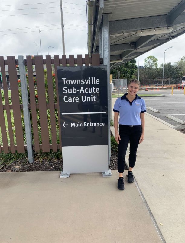 Fourth-year Occupational Therapy student Kady Delle Baite posing, smiling next to the Townsville Sub-Acute Care Unit sign. 
