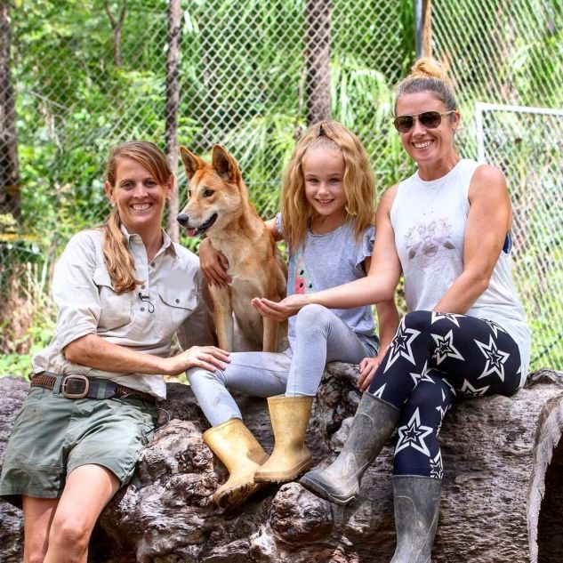 JCU Alumni Shakira Todd smiling and leaning against a log with a tan dingo sitting next to here and a young girl and woman with their hand on the dingo. 