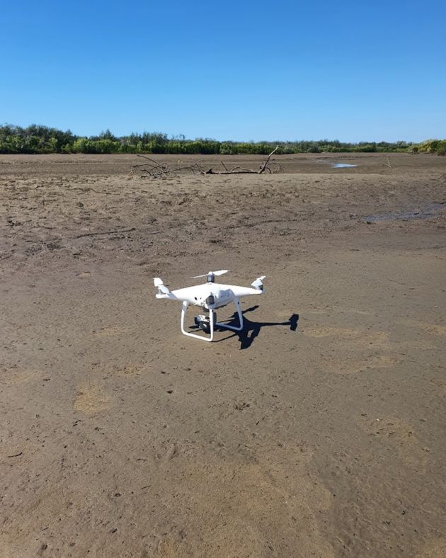A small white drone hovering above sand flats. 
