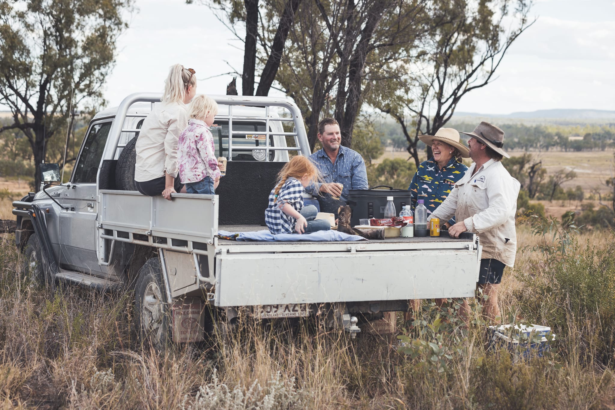 Four adults and two children sit having a picnic in the tray of a ute. 