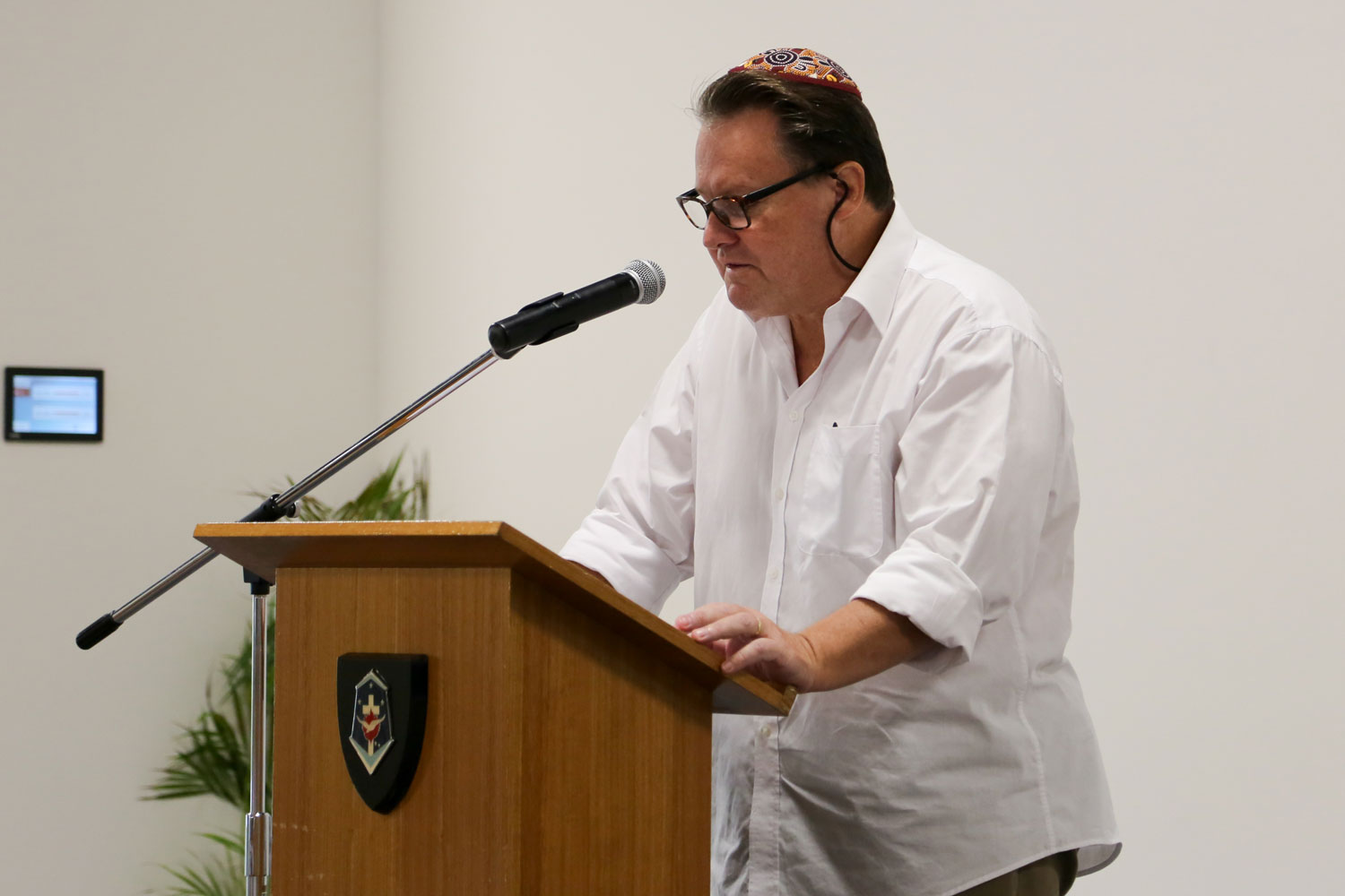 A man in a white shirt wearing a Kippah stands at a lectern talking into a microphone 