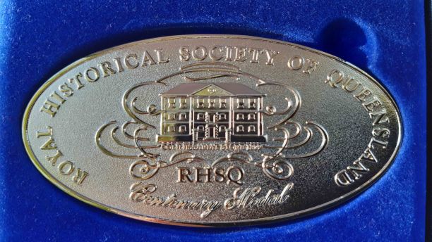 A gold medal laid in a blue box. The medal has Royal Historical Society of Queensland and Centenary Medal engraved on it with the image of a building. 