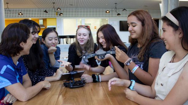 A group of seven girls sitting around a table smiling while they look at a drone. 