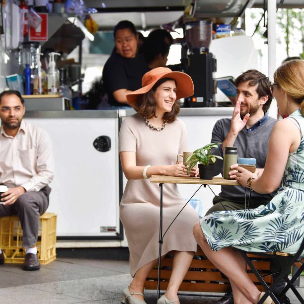 Three people sit at a table drinking coffee in mugs and reusable coffee cups in front of a small coffee van while a man in the background has a single use plastic coffee cup. 
