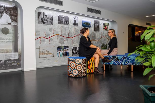 Two students sit together in the Indigenous Education and Research Centre, beside a mural depicting a timeline of Indigenous history. 