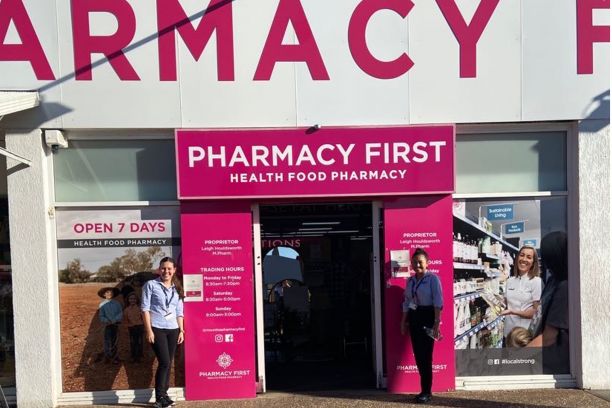 Jai-ann and friend outside the entrance to a pharmacy with large pink letting reading pharmacy above them. 