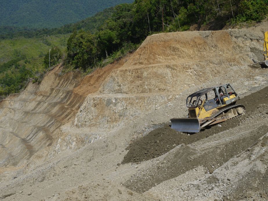 A digger constructing a road in PNG. A large area of dirt has been made amidst a lush green forest. 