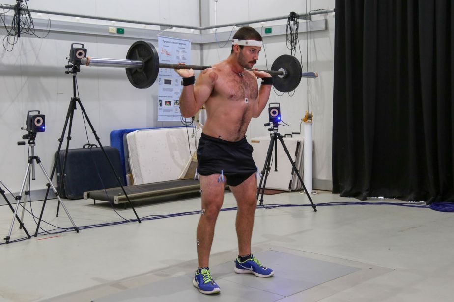 A man wearing small reflectors on his body and on a headband holds a barbell on his shoulders, ready to squat